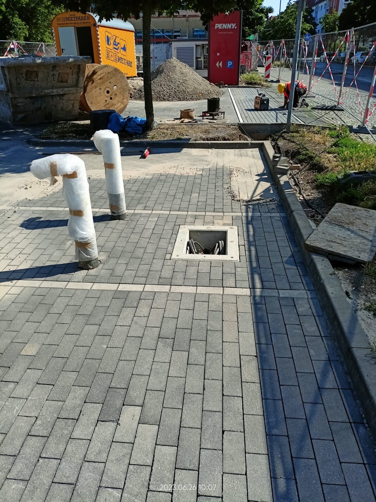 Świeżo ułożona szara kostka brukowa z dwoma zabezpieczonymi słupkami i otwartą studzienką kablową, w tle widoczny teren budowy z kontenerami i stertą żwiru.