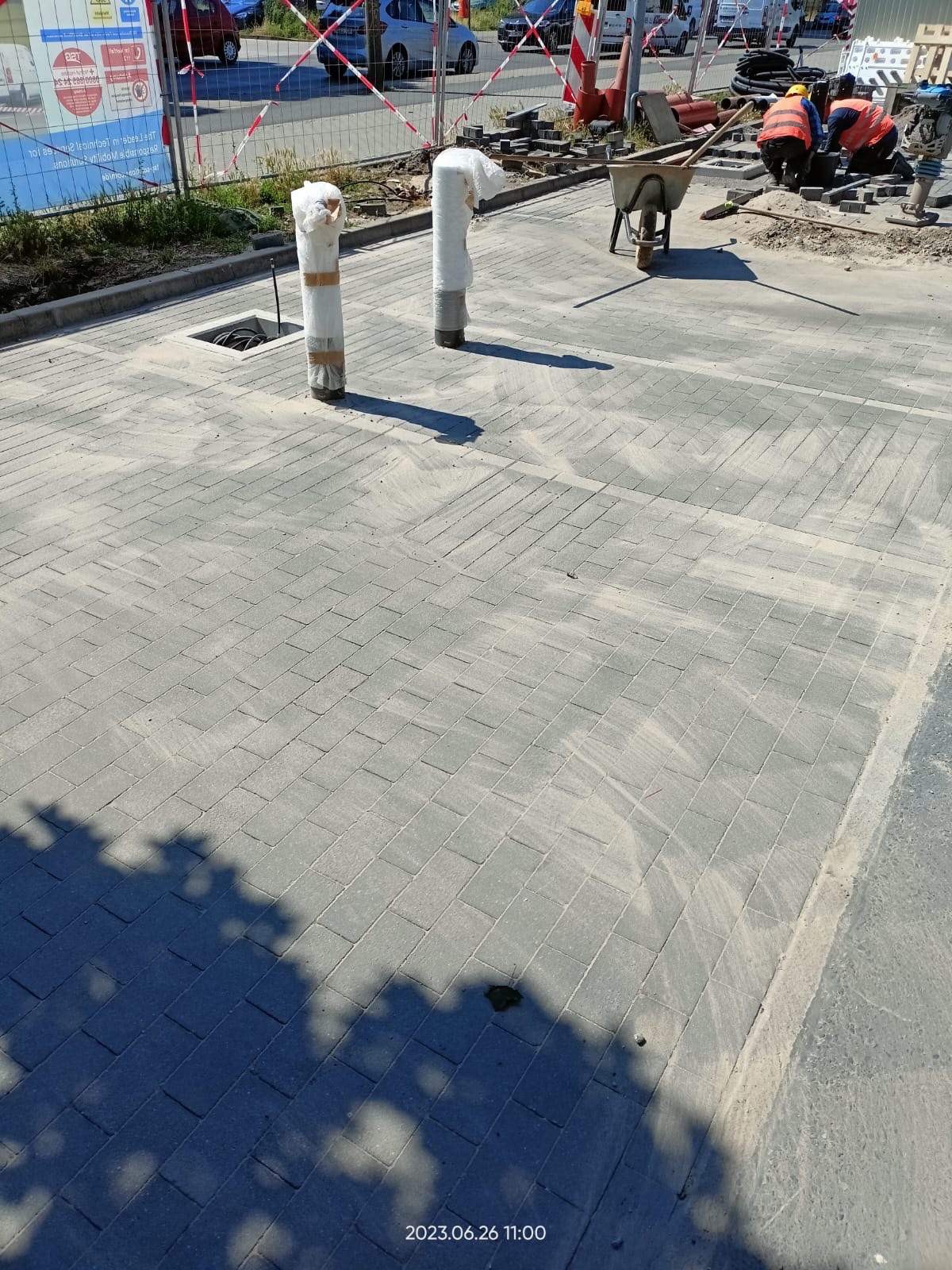 Układanie szarej kostki brukowej na placu, widoczni pracownicy w pomarańczowych kamizelkach, taczka, dwie rury zabezpieczone folią, w tle ogrodzenie i zaparkowane samochody.