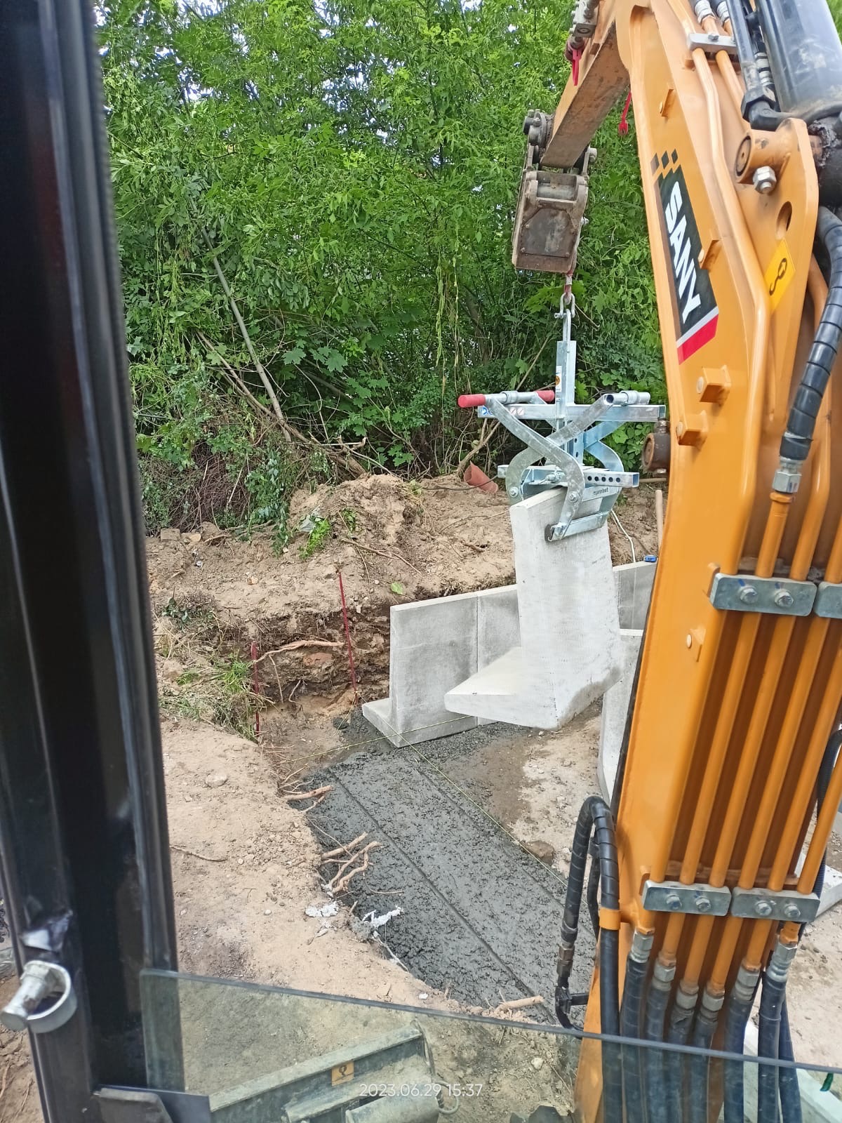 Żuraw przenosi prefabrykowany element betonowy w wykop wypełniony świeżym betonem, widok z kabiny operatora.