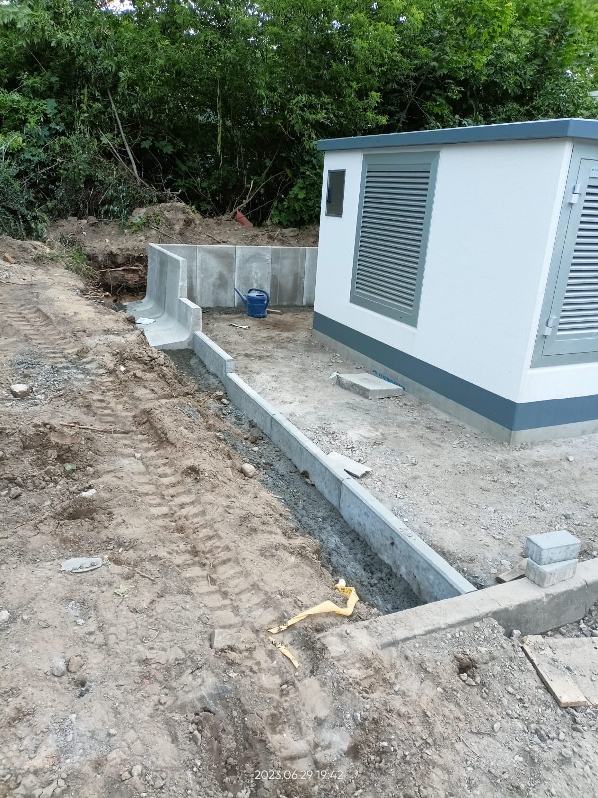 Budowa fundamentów pod budynek gospodarczy z betonowymi obrzeżami i śladami opon na ziemi, widoczna niebieska konewka i fragment lasu w tle.