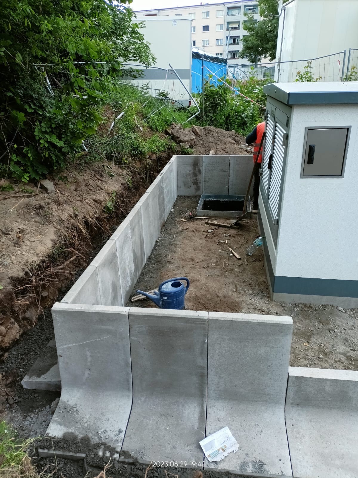 Budowa betonowego fundamentu pod budynek gospodarczy z widoczną niebieską konewką i pracownikiem w pomarańczowej kamizelce.