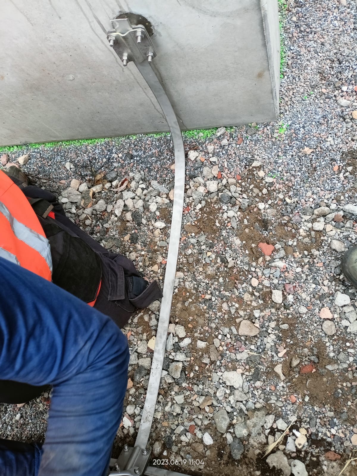 Metalowa listwa uziemiająca przymocowana do betonowej konstrukcji z widocznymi śrubami i elementami mocującymi, leżąca na żwirowym podłożu z fragmentem odzieży roboczej i buta.