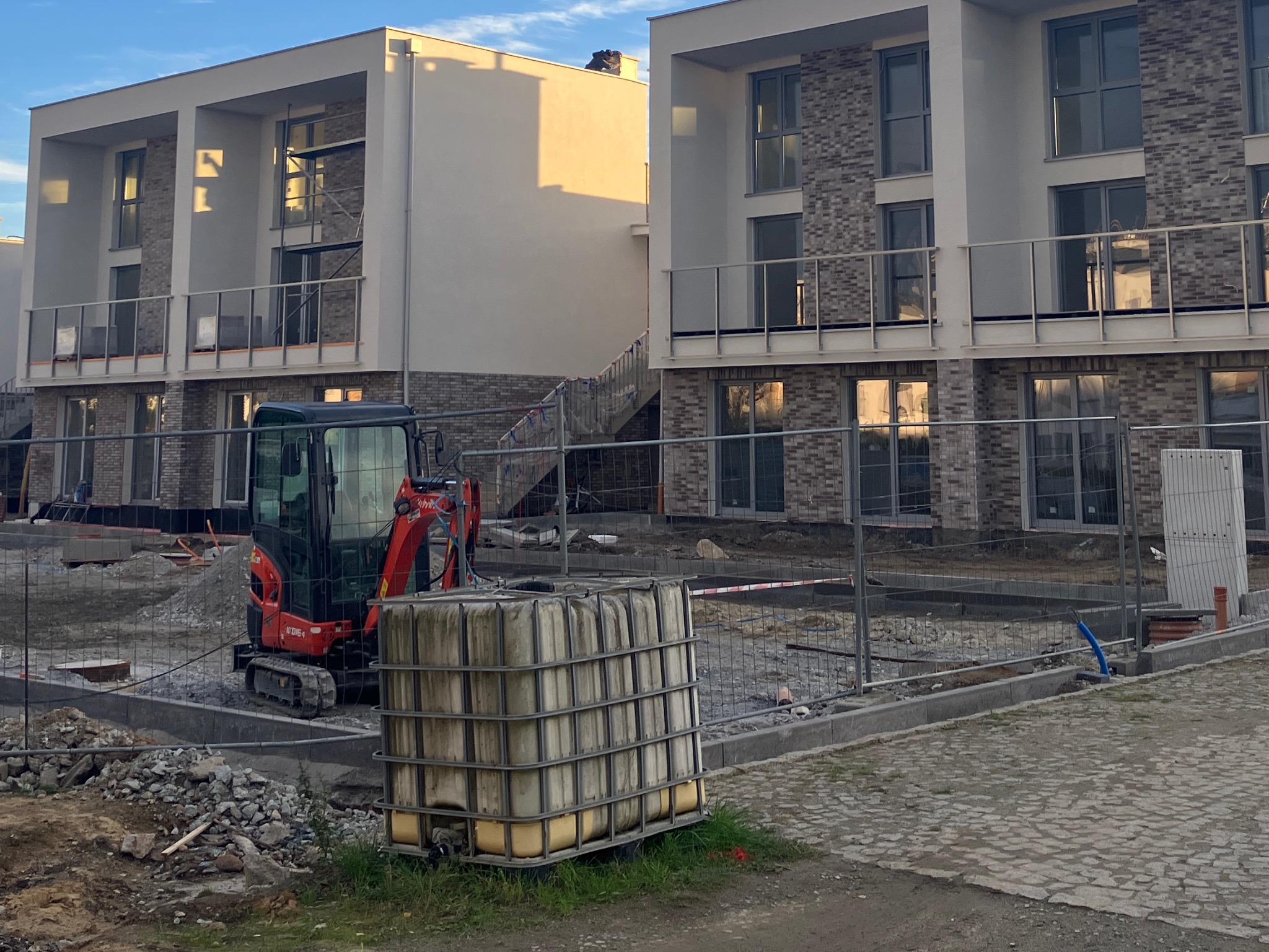 Nowo budowane, dwupiętrowe budynki mieszkalne z ceglaną elewacją i balkonami, widoczne przez ogrodzenie placu budowy, z mini koparką i zbiornikiem na wodę w tle.