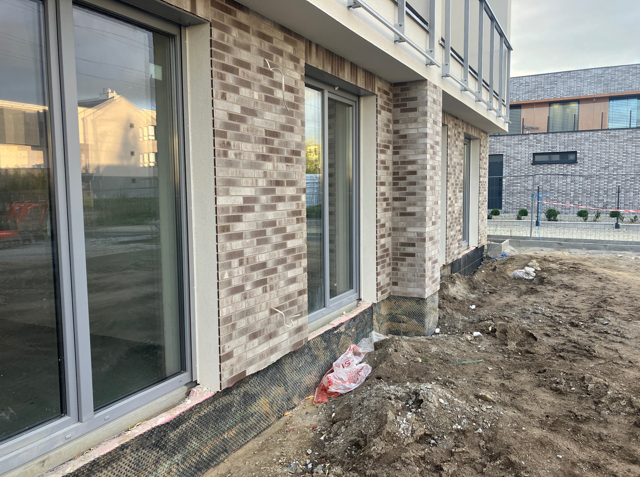 Nowoczesny budynek w trakcie wykończenia elewacji z cegły klinkierowej, widoczne okna z szarymi ramami, grunt wokół budynku niezagospodarowany, folia ochronna.