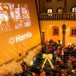 Wewnętrzny dziedziniec z widokiem na zgromadzony tłum ludzi, oświetlony girlandami światełek i dekoracjami świątecznymi, z projekcją logo 'Homla' na ścianie w tle.