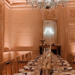 Plannerki sp. z o.o. - Uroczysta sala balowa z długim stołem nakrytym białym obrusem, złotymi krzesłami, kryształowym żyrandolem i świecami, odbijającymi się w lustrze nad kominkiem.
