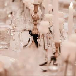 Plannerki sp. z o.o. - Elegancko nakryty stół z kryształowymi kieliszkami, złotym świecznikiem z czarną wstążką, białymi różami w wazonach i płonącą świecą, utrzymany w ciepłej tonacji.