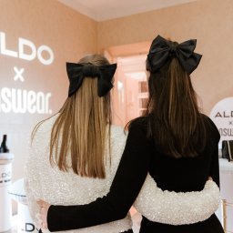 Plannerki sp. z o.o. - Dwie kobiety, tyłem, obejmują się na tle ściany z logo Aldo x Unswear, obie mają czarne kokardy we włosach, jedna w cekinowej kurtce, druga w czarnej sukience.