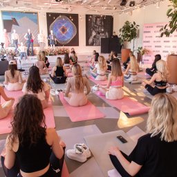 Plannerki sp. z o.o. - Grupa kobiet siedzących na różowych matach do jogi w jasnym pomieszczeniu z dekoracjami i logo GUESS, prowadzący zajęcia na podwyższeniu.