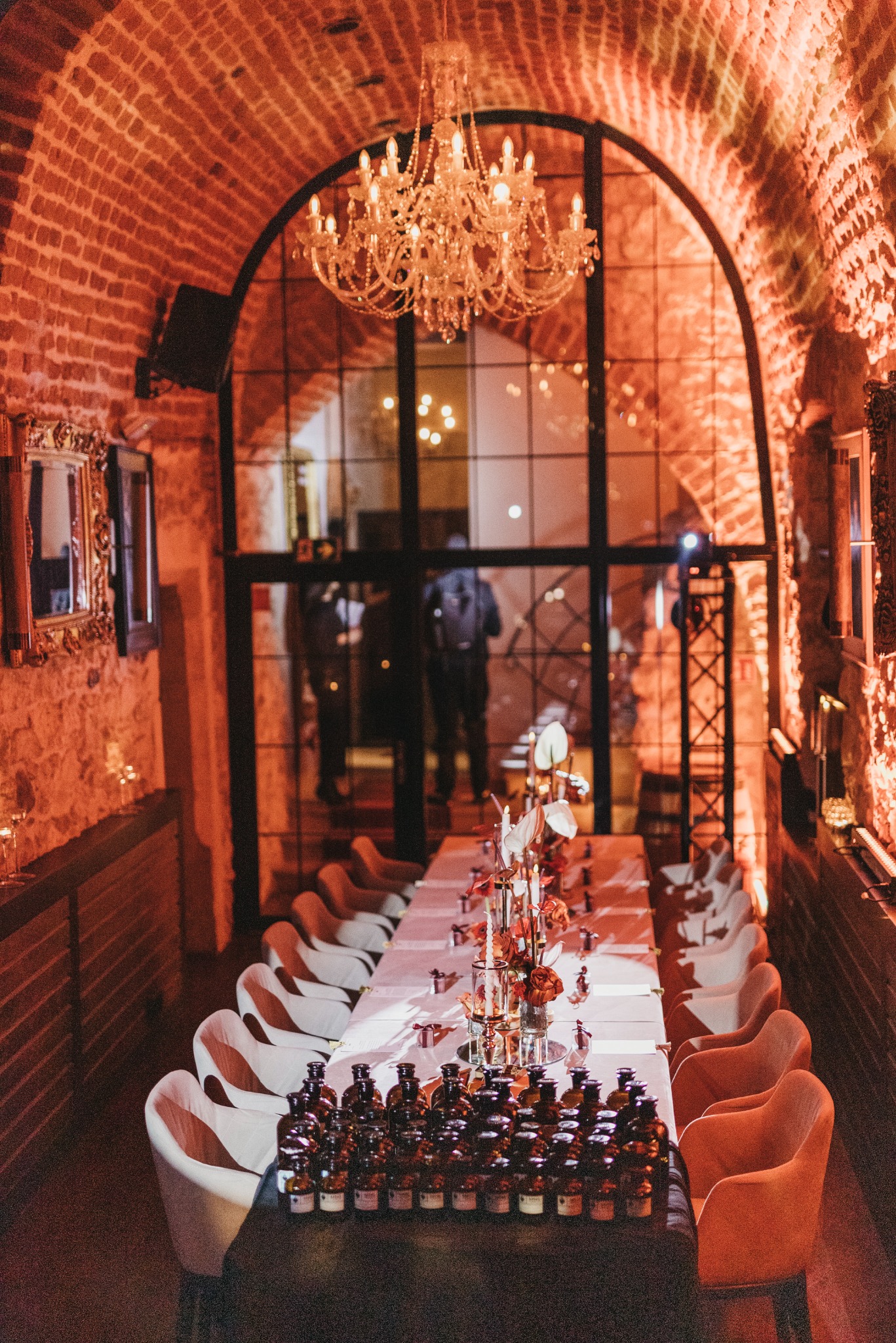 Uroczysta kolacja firmowa w ceglanym wnętrzu w Krakowie, długi stół z białym obrusem, dekoracje kwiatowe i świeczniki, rzędy małych buteleczek, krzesła w odcieniach bieli i pomarańczy, kryształowy...