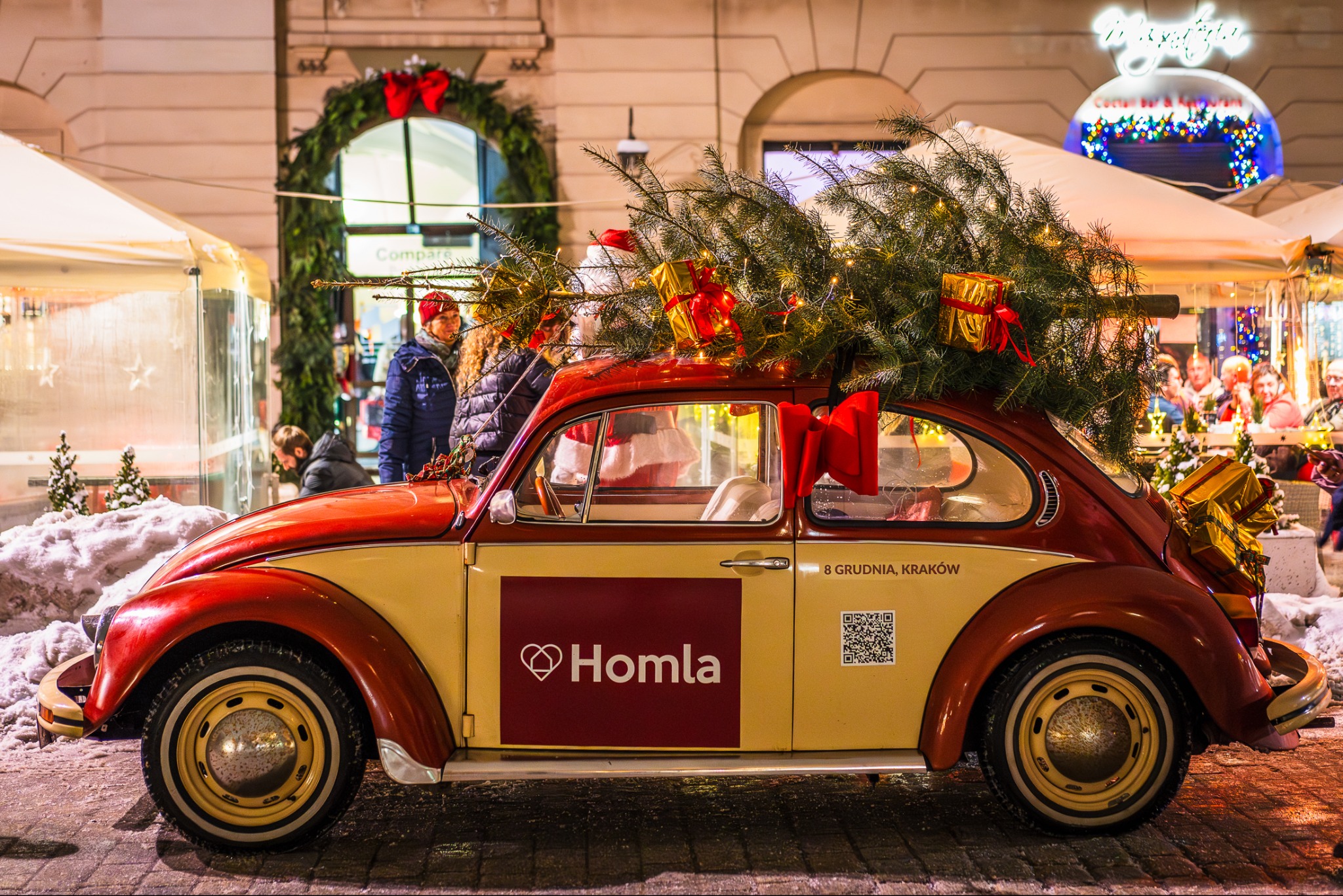 Czerwono-beżowy Volkswagen Garbus z choinką i prezentami na dachu, zaparkowany na brukowanej ulicy, z logo Homla na drzwiach i adresem '8 Grudnia, Kraków', w tle świątecznie udekorowane budynki...