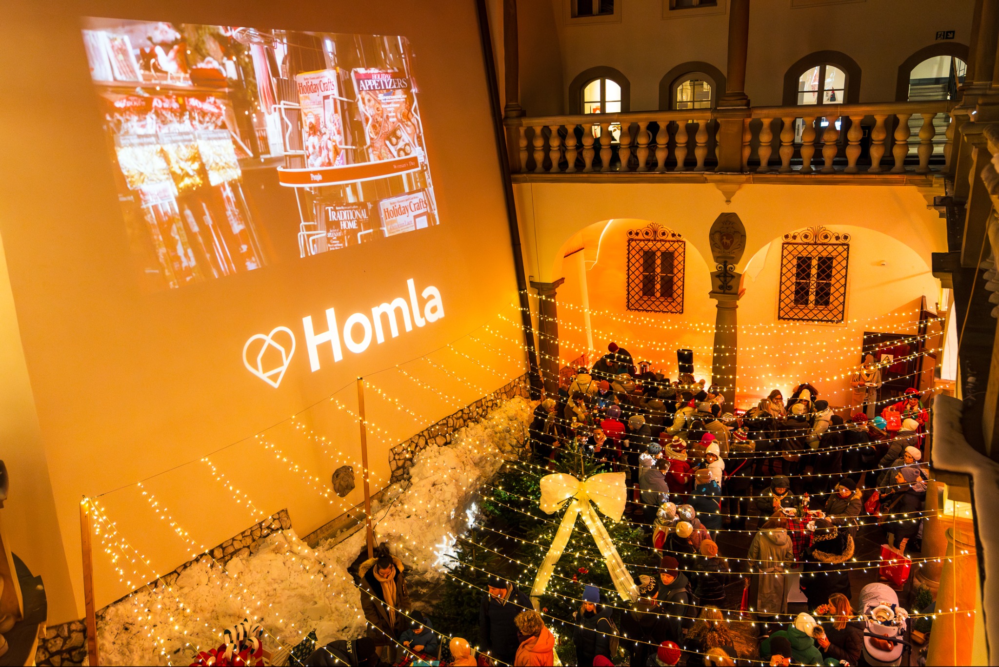 Wewnętrzny dziedziniec z widokiem na zgromadzony tłum ludzi, oświetlony girlandami światełek i dekoracjami świątecznymi, z projekcją logo 'Homla' na ścianie w tle.
