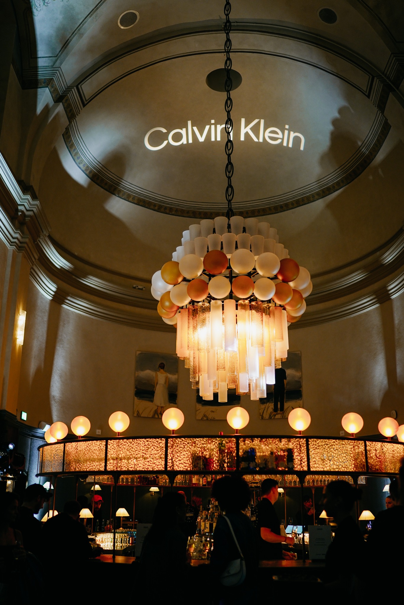 Wnętrze baru z oświetleniem punktowym, wiszącym żyrandolem z okrągłych i prostokątnych elementów oraz projekcją logo Calvin Klein na suficie kopułowym.