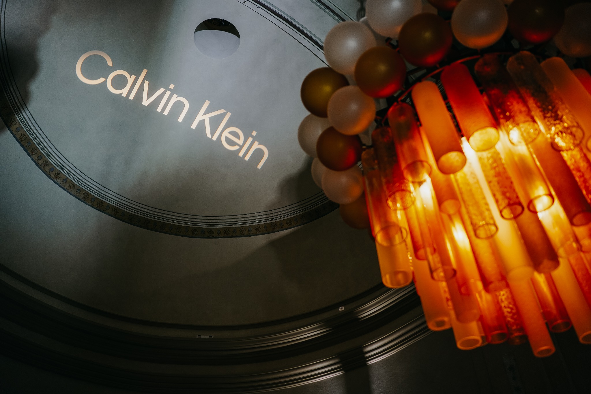 Uroczyste wnętrze z dekoracją balonową i pomarańczowym żyrandolem, na tle sufitu z napisem 'Calvin Klein'.