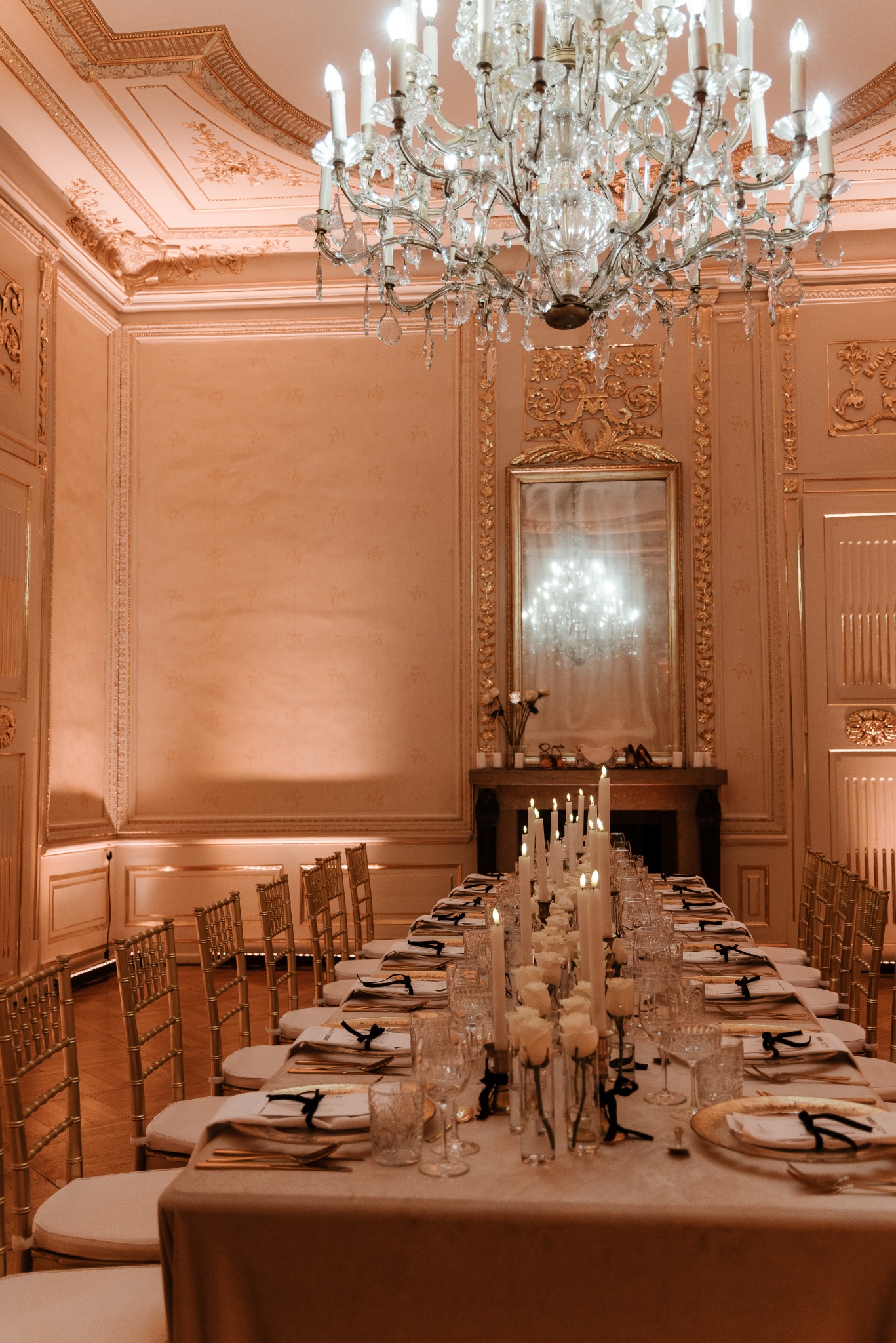Uroczysta sala balowa z długim stołem nakrytym białym obrusem, złotymi krzesłami, kryształowym żyrandolem i świecami, odbijającymi się w lustrze nad kominkiem.