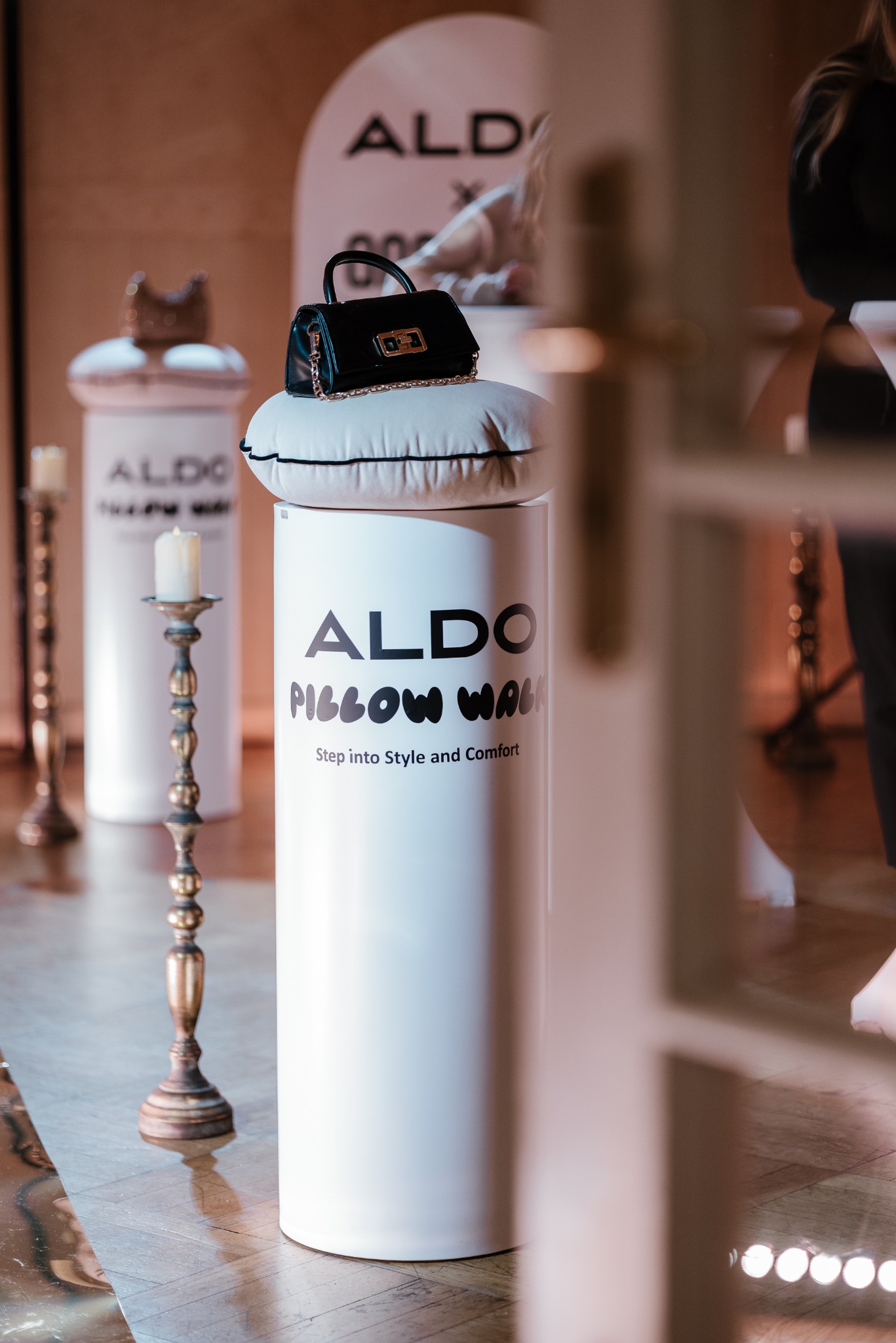 Czarna, lakierowana torebka z łańcuszkiem na białej poduszce, umieszczona na białym postumencie z napisem 'ALDO Pillow Walk', w tle dekoracje i świeczniki, widok przez framugę drzwi.