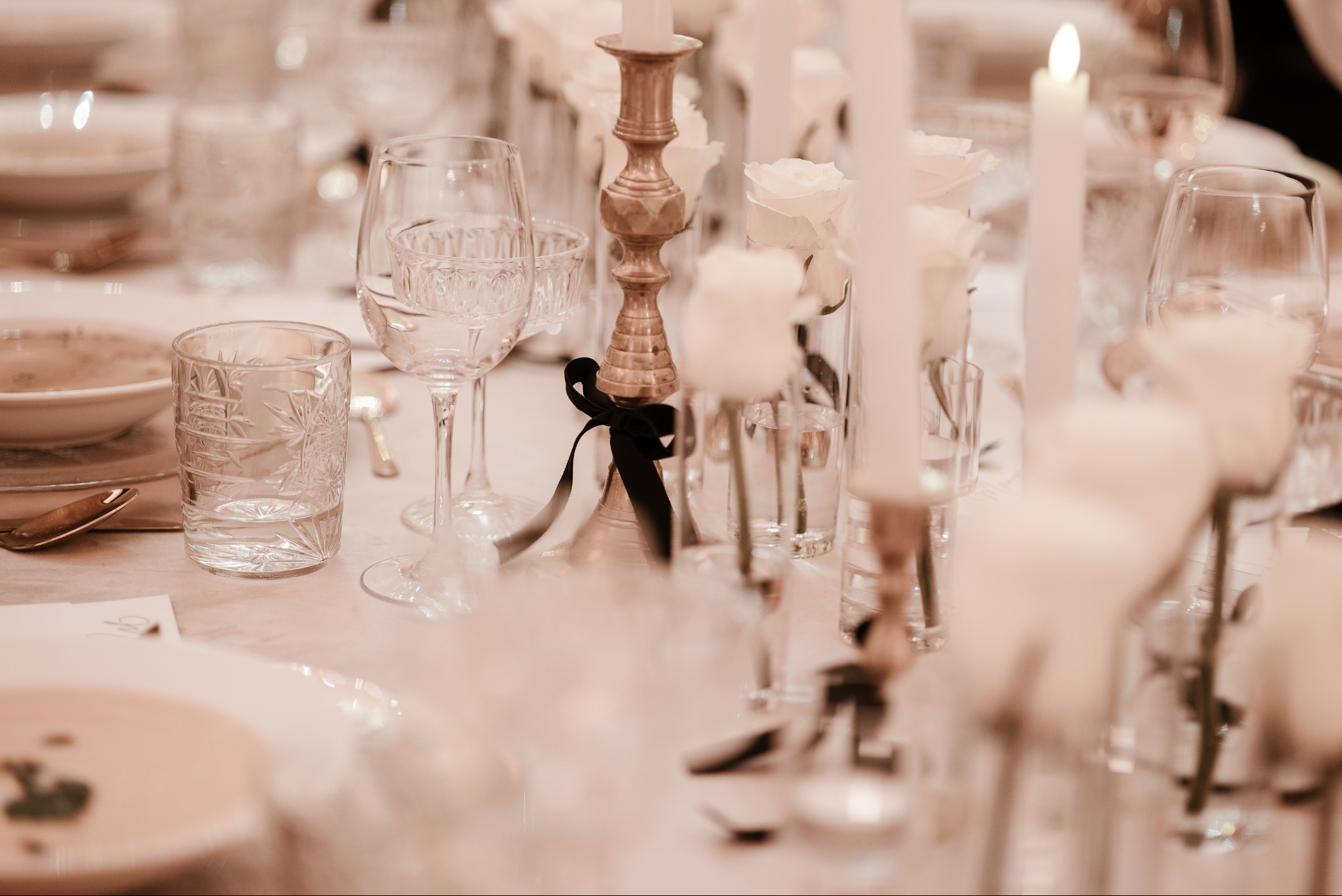 Elegancko nakryty stół z kryształowymi kieliszkami, złotym świecznikiem z czarną wstążką, białymi różami w wazonach i płonącą świecą, utrzymany w ciepłej tonacji.
