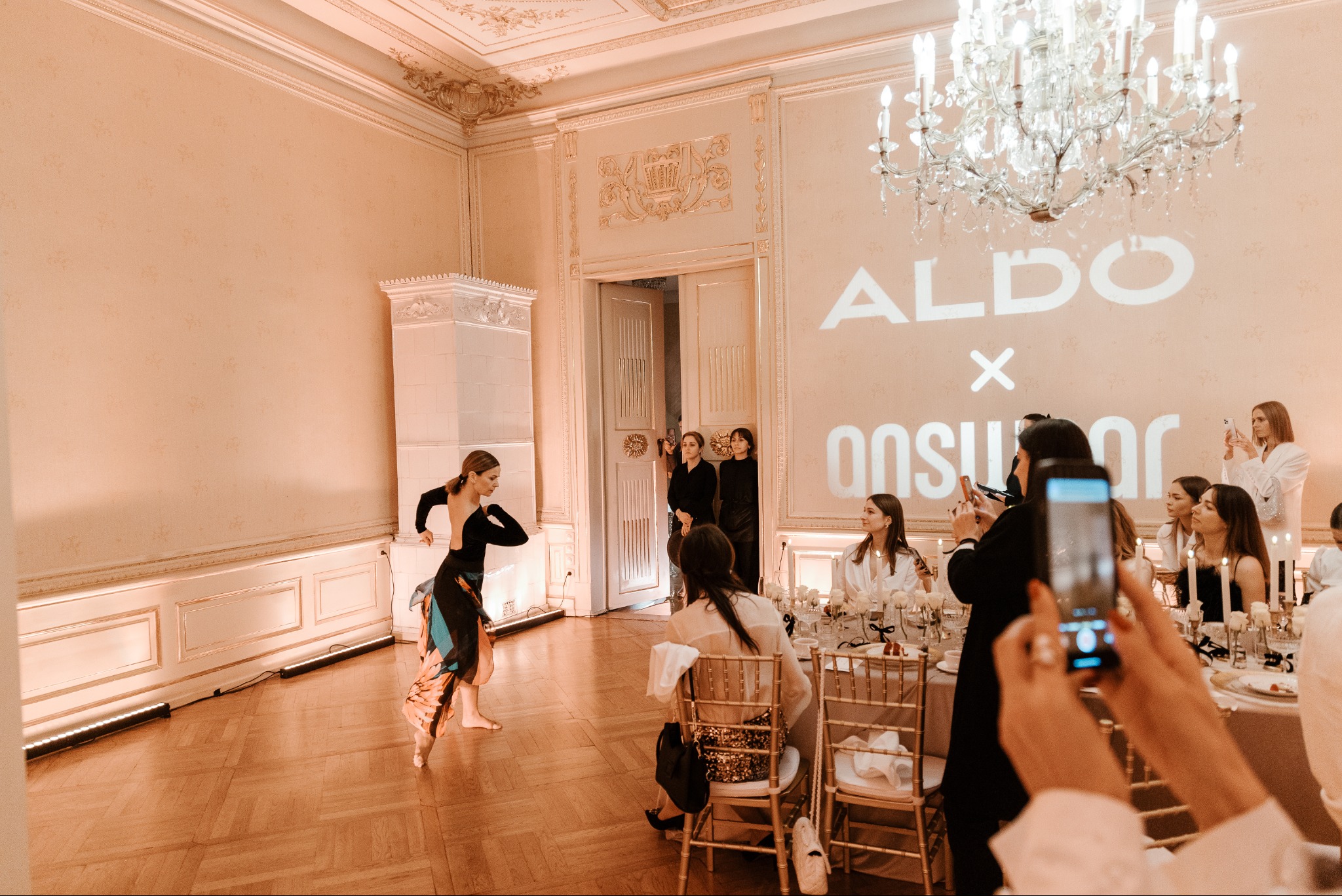 Tancerka w ruchu na parkiecie, goście przy stołach obserwują i filmują, na ścianie wyświetlane logo ALDO x ANSWEAR, kryształowy żyrandol, eleganckie wnętrze.