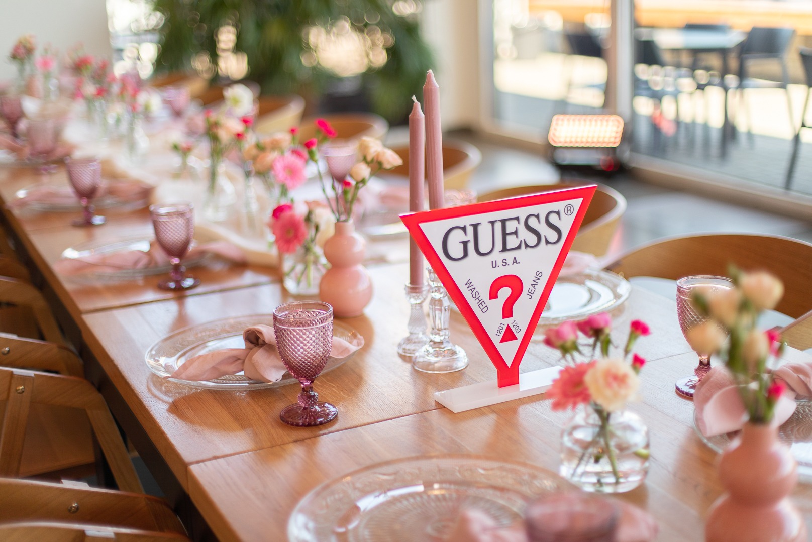 Uroczysty stół z dekoracjami w odcieniach różu: szklane naczynia, kwiaty w małych wazonach, wysokie świece i kartonowe logo Guess jako element dekoracyjny.