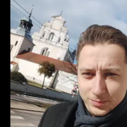 Autoportret mężczyzny w szarym szaliku i czarnym płaszczu na tle kościoła z wieżami, z widocznym przejściem dla pieszych i znakami drogowymi.