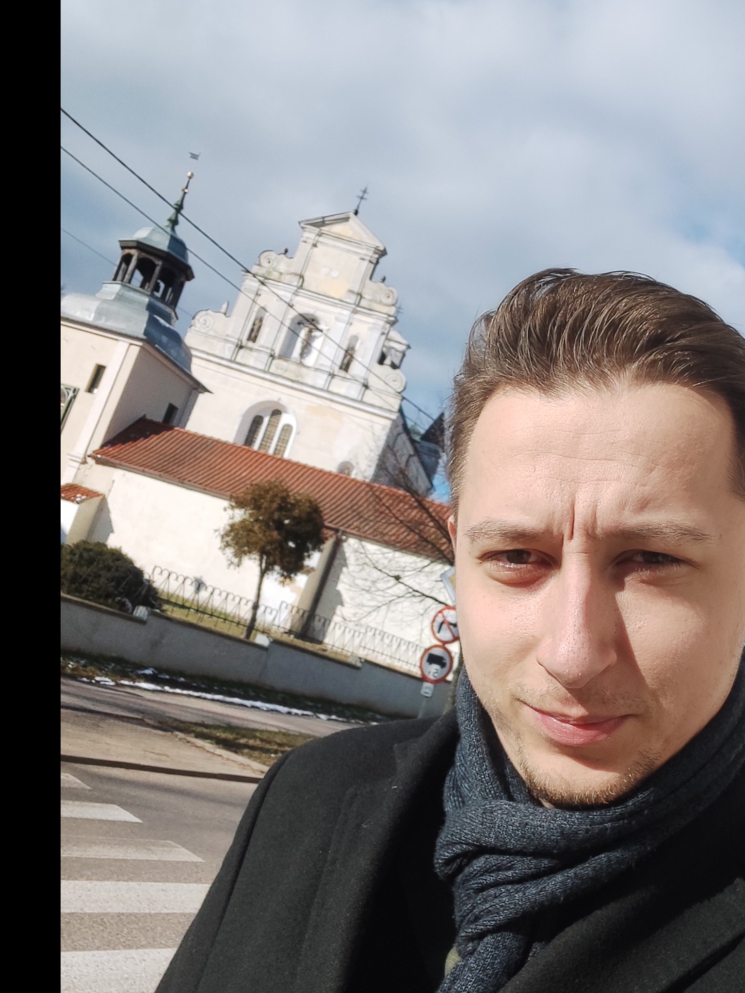 Autoportret mężczyzny w szarym szaliku i czarnym płaszczu na tle kościoła z wieżami, z widocznym przejściem dla pieszych i znakami drogowymi.