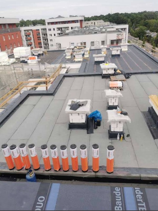 Prace na płaskim dachu budynku wielorodzinnego w Chojnowie, widoczne materiały izolacyjne w pomarańczowych tubach, elementy wentylacyjne z cegły oraz częściowo położona papa.