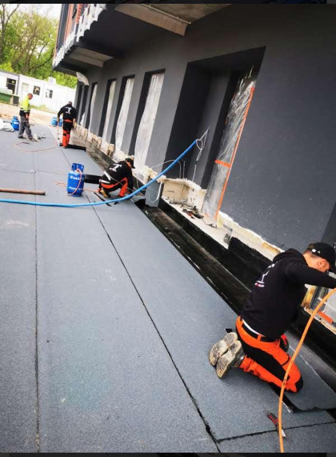 Pracownicy w pomarańczowych spodniach układają papę termozgrzewalną na dachu budynku z czarną elewacją, widoczne butle gazowe i narzędzia.