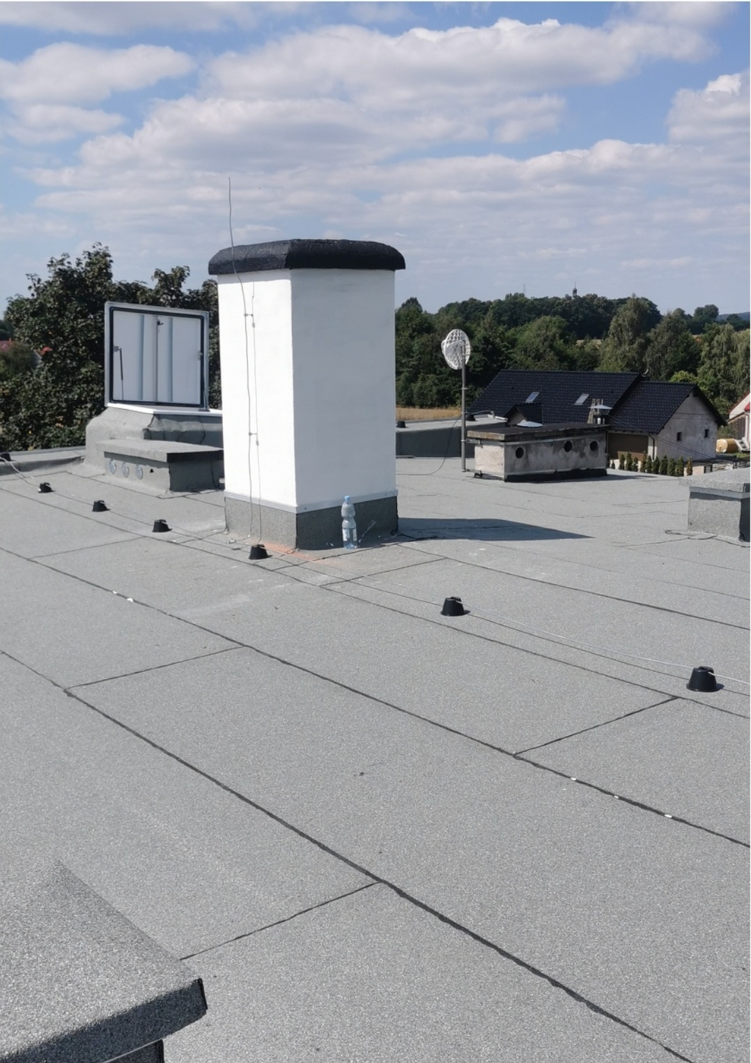 Szary, płaski dach pokryty papą termozgrzewalną z widocznym kominem, wyłazem dachowym i elementami wentylacyjnymi na tle błękitnego nieba.