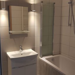 Jacek - Wykończona łazienka: biała umywalka z szafką, lustro z oświetleniem, wanna z parawanem i panelami imitującymi drewno, płytki w odcieniach szarości.