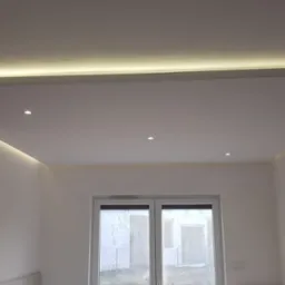 Podwieszany sufit z oświetleniem LED, trzy punktowe lampy sufitowe, białe ściany, widoczne okno balkonowe, drabina oparta o ścianę, surowy stan wykończenia.