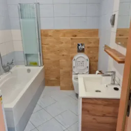 Ukończona łazienka z wanną z parawanem, toaletą i umywalką z szafką w odcieniach bieli, brązu i szarości, widok zza otwartych drzwi.