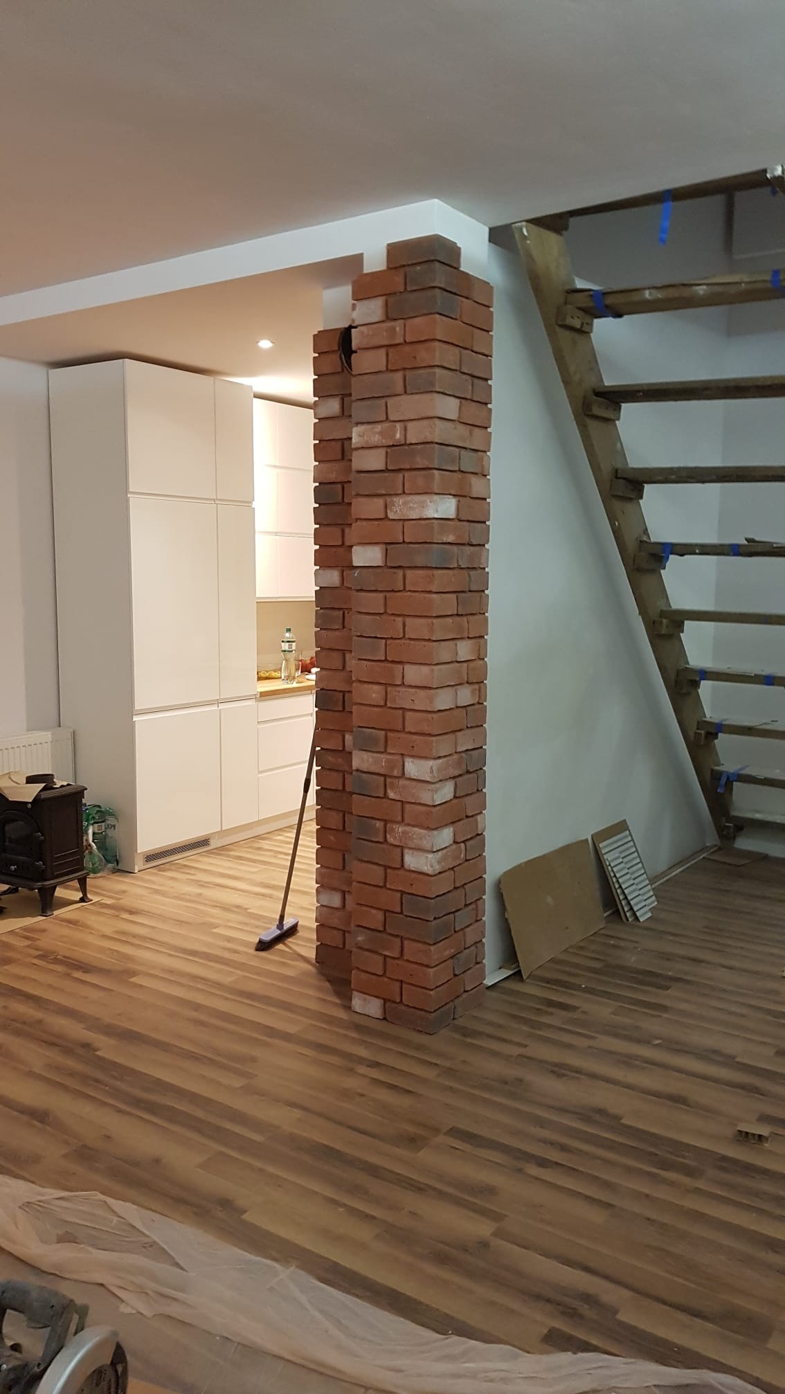 Murowana kolumna z cegły klinkierowej w surowym wnętrzu, widoczne drewniane schody, podłoga z paneli i białe meble kuchenne w tle, mop oparty o kolumnę.