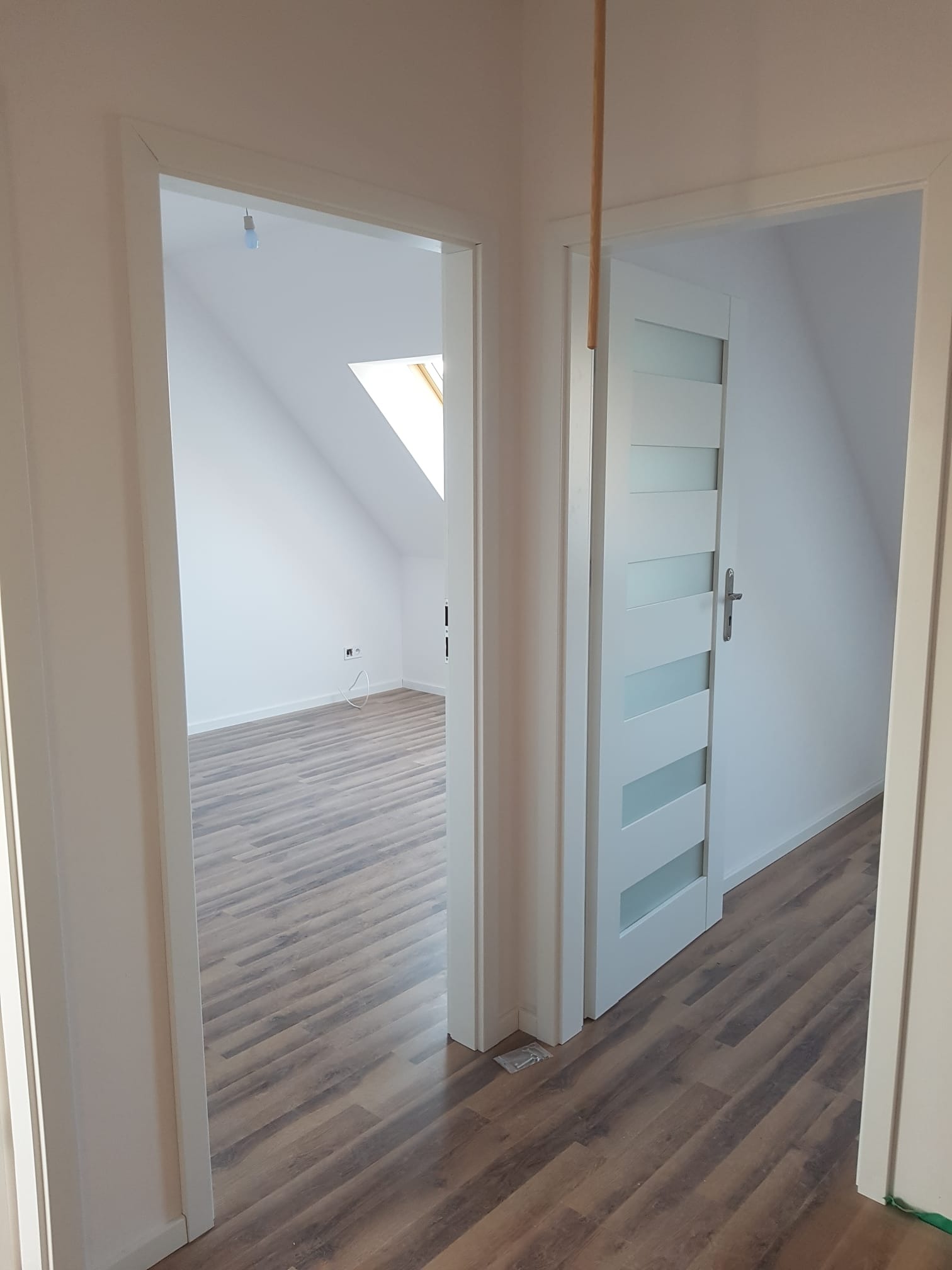 Widok na wykończone poddasze z panelami podłogowymi i zamontowanymi białymi drzwiami z przeszkleniami, widoczne przez dwa otwarte przejścia.