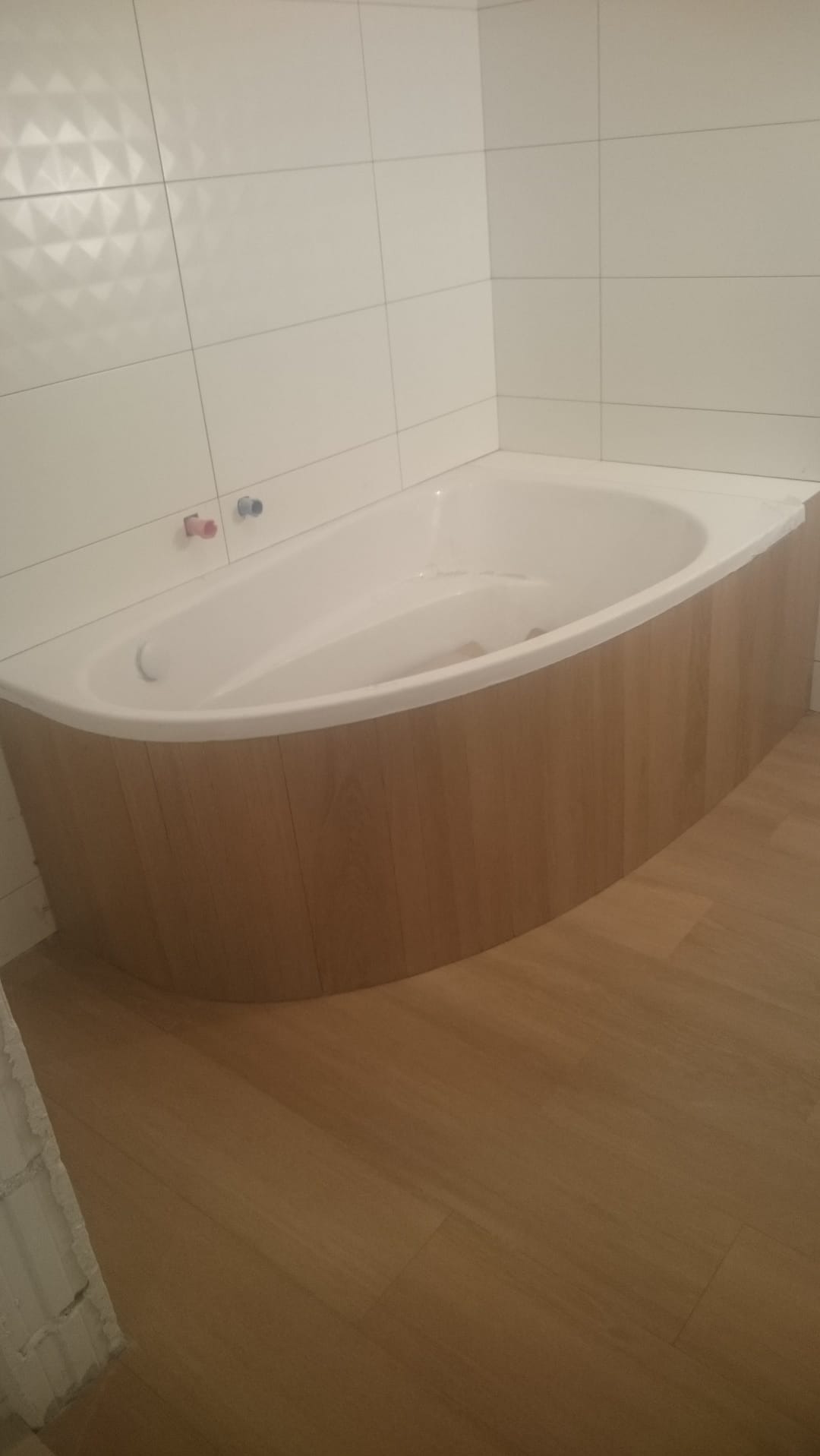 Biała wanna obudowana panelami imitującymi drewno, białe kwadratowe płytki na ścianie, podłoga wyłożona panelami podłogowymi w kolorze jasnego drewna, widoczne zawory do baterii.