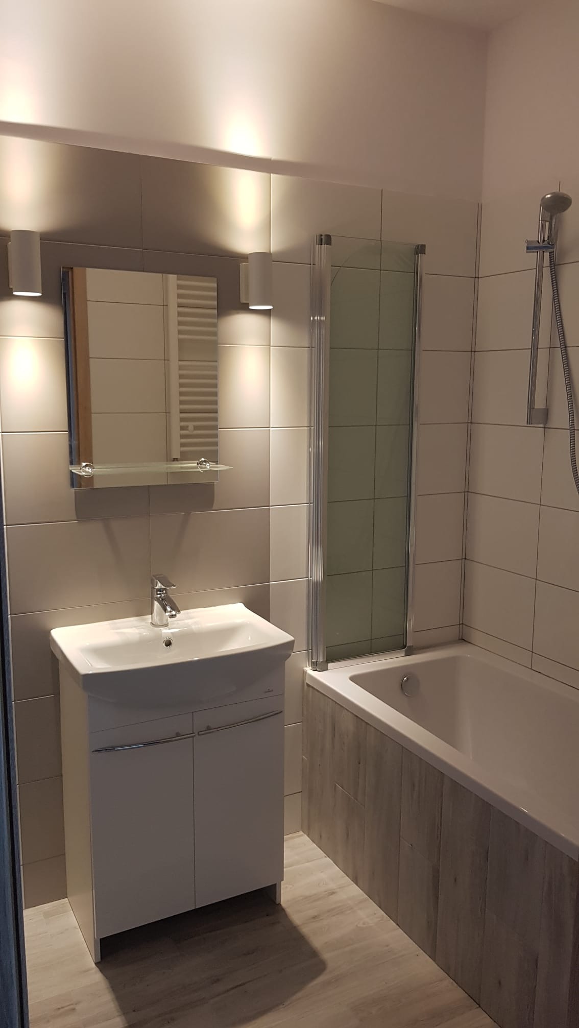 Wykończona łazienka: biała umywalka z szafką, lustro z oświetleniem, wanna z parawanem i panelami imitującymi drewno, płytki w odcieniach szarości.