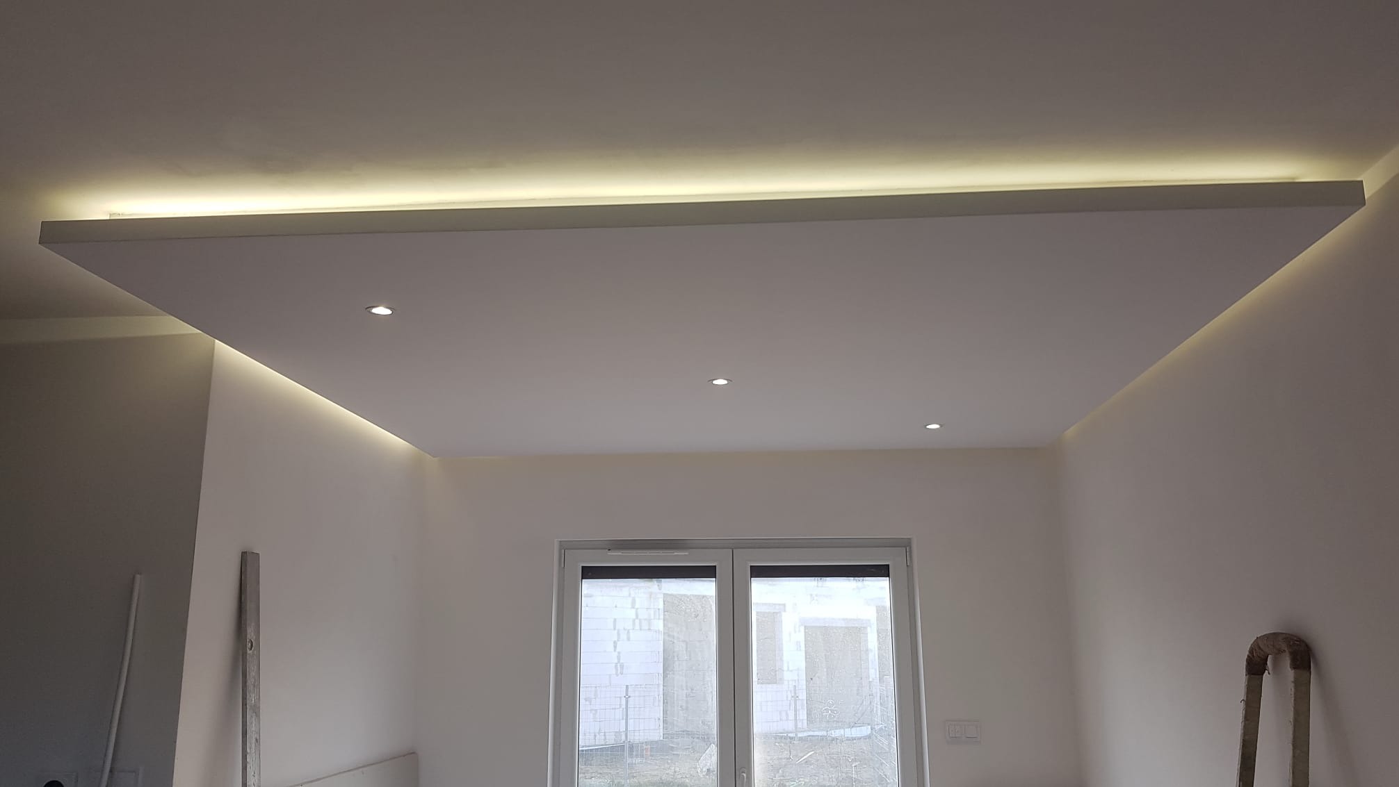 Podwieszany sufit z oświetleniem LED, trzy punktowe lampy sufitowe, białe ściany, widoczne okno balkonowe, drabina oparta o ścianę, surowy stan wykończenia.
