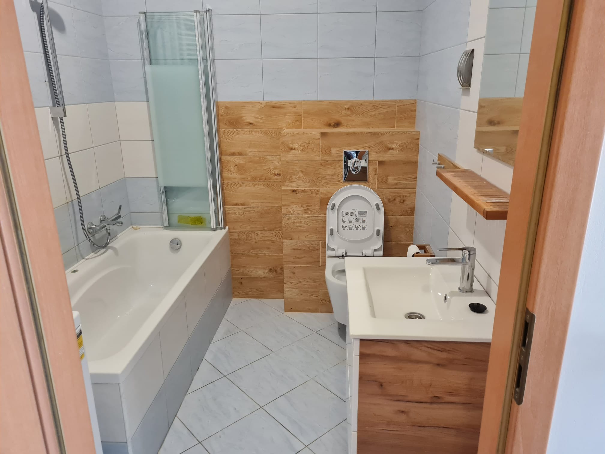 Ukończona łazienka z wanną z parawanem, toaletą i umywalką z szafką w odcieniach bieli, brązu i szarości, widok zza otwartych drzwi.