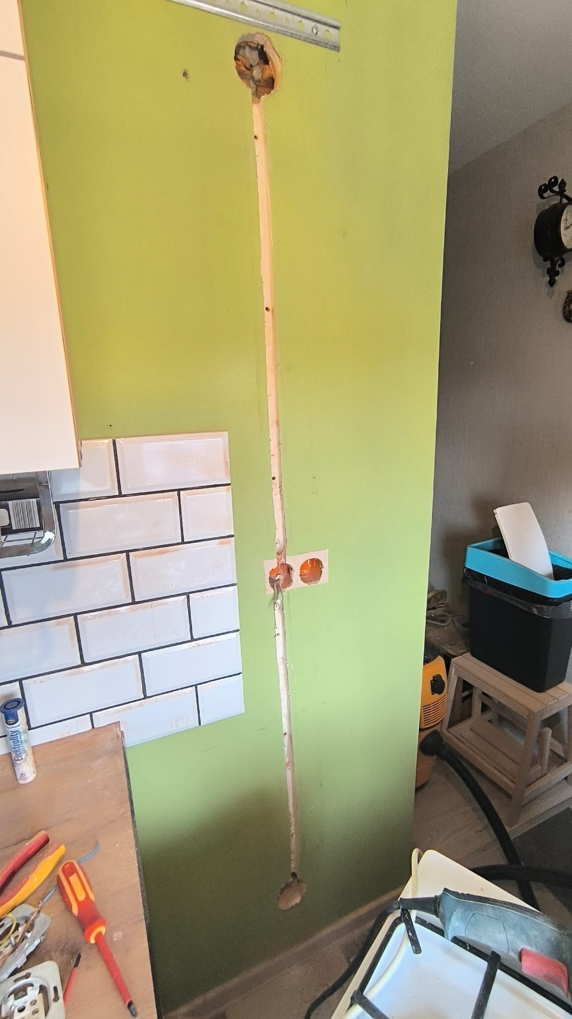 Wnęka w zielonej ścianie z wyfrezowanym kanałem na przewody i otworem na gniazdko, obok fragmentu ściany z białymi płytkami, widoczne narzędzia na blacie, w tle fragment wyposażenia kuchni.