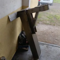 Surowa, spawana metalowa konstrukcja wspornika z przymocowanym elementem do ściany, ustawiona w warsztacie obok samochodu dostawczego i pudełka narzędziowego.