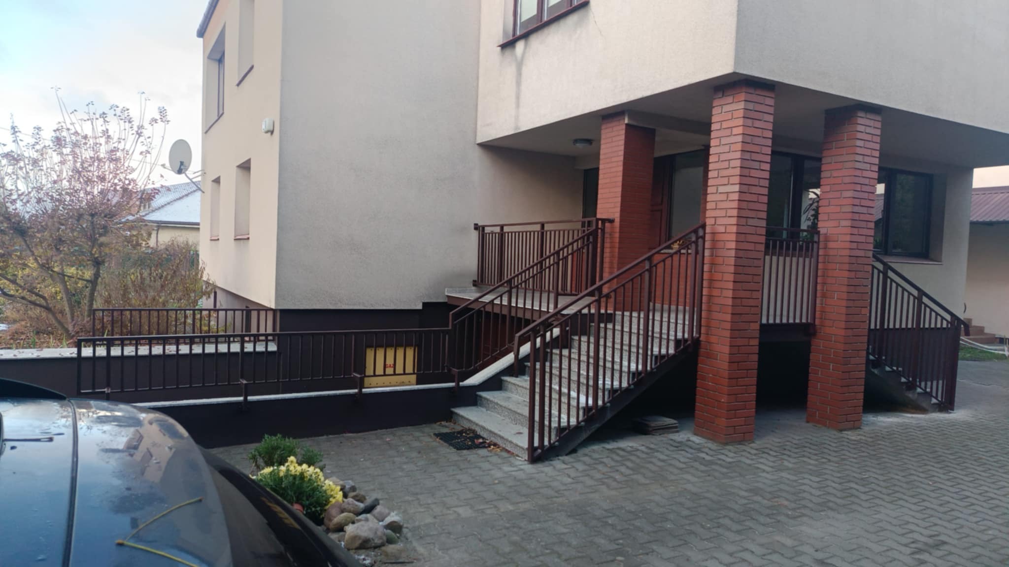 Zewnętrzne schody wejściowe do budynku z brązowymi metalowymi poręczami i balustradami, wsparte na ceglanych filarach, widok z poziomu chodnika.