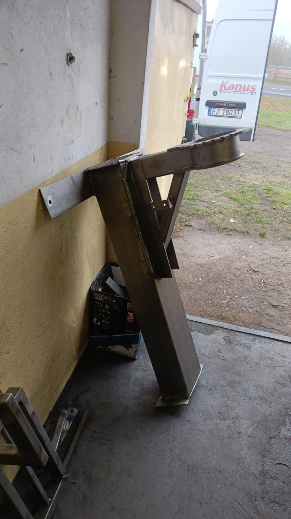 Surowa, spawana metalowa konstrukcja wspornika z przymocowanym elementem do ściany, ustawiona w warsztacie obok samochodu dostawczego i pudełka narzędziowego.