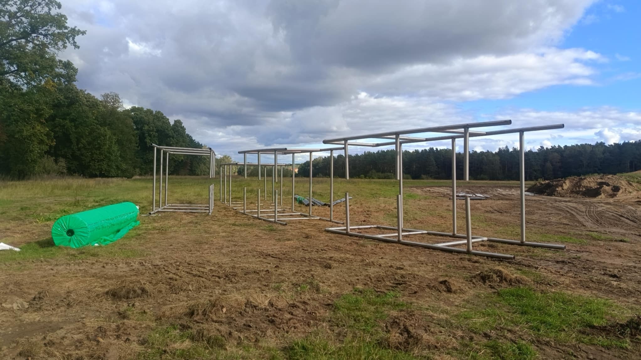 Konstrukcja metalowa pod ogrodzenie, widoczne ramy z rur na nierównym terenie z rolką zielonej siatki w tle, etap montażu.