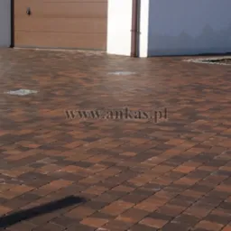 Ułożona, brązowo-czerwona kostka brukowa na podjeździe przed budynkiem z brązową bramą garażową i rynną spustową.