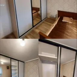 Biała szafa wnękowa z czarnymi obramowaniami drzwi przesuwnych i lustrami, odbijająca wnętrze sypialni z łóżkiem i tapetą w kwiatowy wzór.