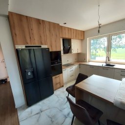 Hubert Czarkowski - Jasna kuchnia z drewnianymi i białymi szafkami, czarną lodówką i marmurową podłogą. Widok na okno z zielenią w tle. Nowoczesny design.