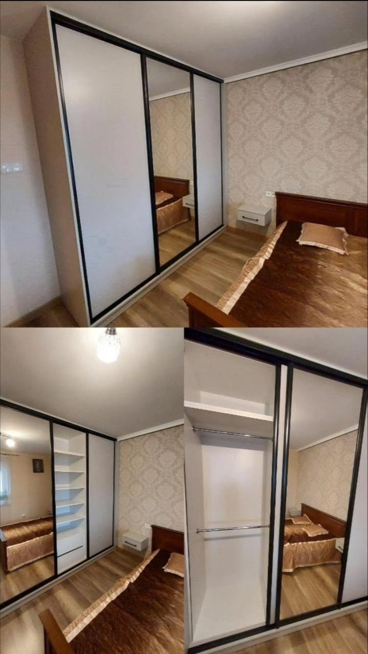 Biała szafa wnękowa z czarnymi obramowaniami drzwi przesuwnych i lustrami, odbijająca wnętrze sypialni z łóżkiem i tapetą w kwiatowy wzór.