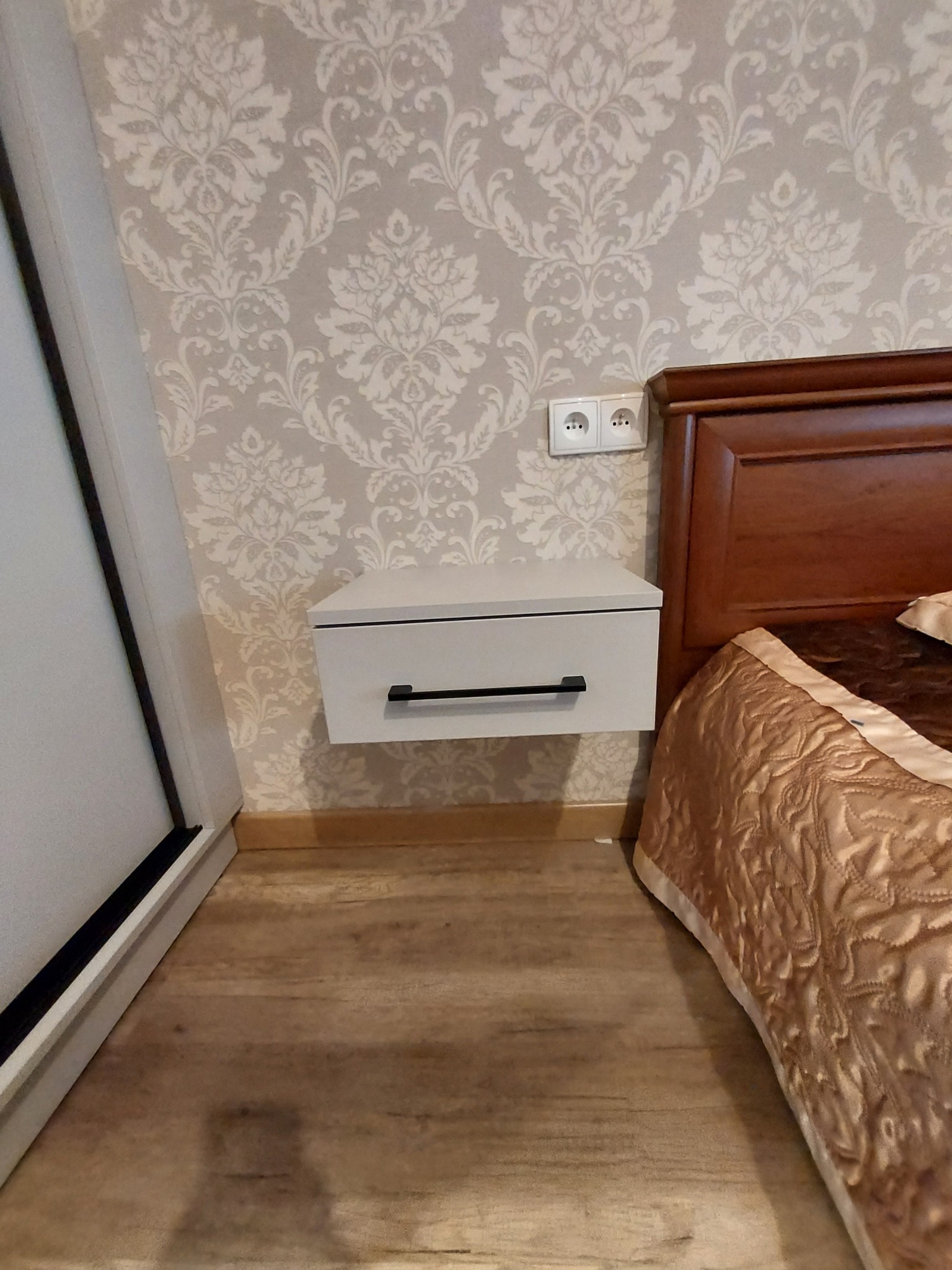 Biała szafka nocna z czarnym uchwytem, zawieszona na ścianie z wzorzystą tapetą, obok fragmentu łóżka z brązową narzutą. Widoczne gniazdko elektryczne.