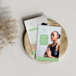 Dwie wizytówki studia jogi 'lõgi' na drewnianej podstawce, z dekoracyjną gałązką trawy w tle. Jedna wizytówka prezentuje ofertę zajęć, druga przedstawia kobietę w pozycji medytacyjnej.