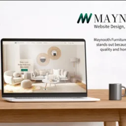 Laptop wyświetla stronę internetową Maynooth Furniture z aranżacją wnętrza w stylu skandynawskim, na biurku obok lampka i kubek.