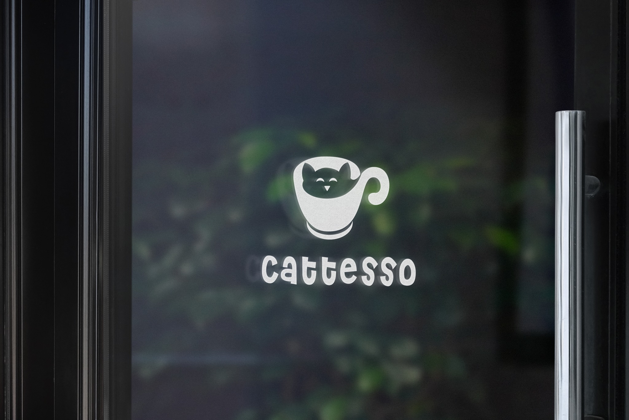 Logo 'Cattesso' na szklanych drzwiach wejściowych, przedstawiające kota w filiżance, z zielonymi roślinami w tle.